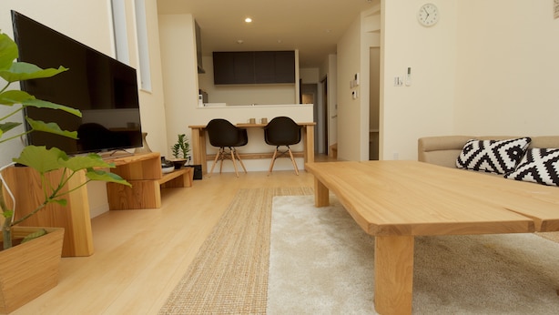 Ett modernt vardagsrum med ett soffbord i trä, två svarta stolar, en beige soffa med mönstrade kuddar, en platt-TV och ett köksområde i bakgrunden.