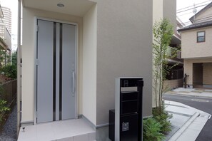 A modern building with a sliding door, a small outdoor area with a planter, and a black structure that could be a utility box or equipment. - Nishi-Sugamo Guest House. 2min to Nishi-Sugamo Sta. 13min to Ikebukuro Sta. (Tokyo)