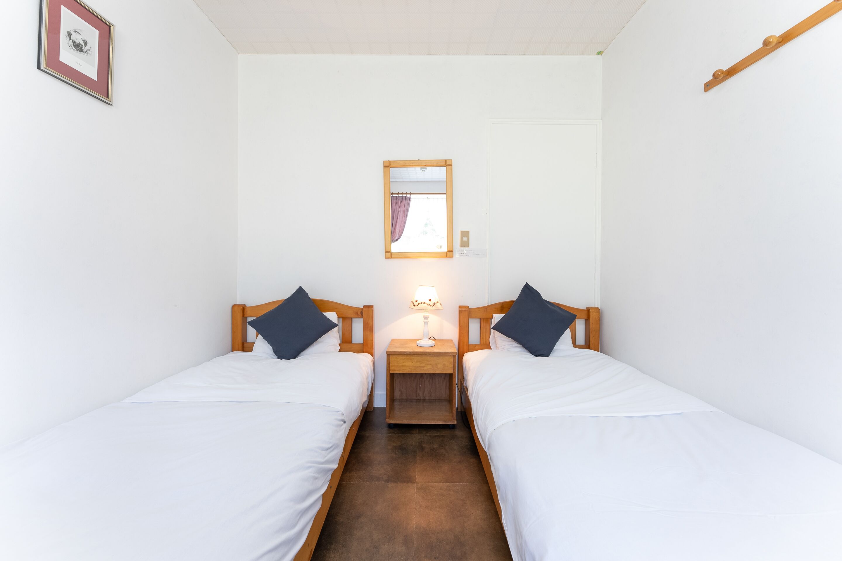 Une chambre avec deux lits simples, une table de chevet avec une lampe, un tableau encadré au mur et une poutre en bois au plafond.