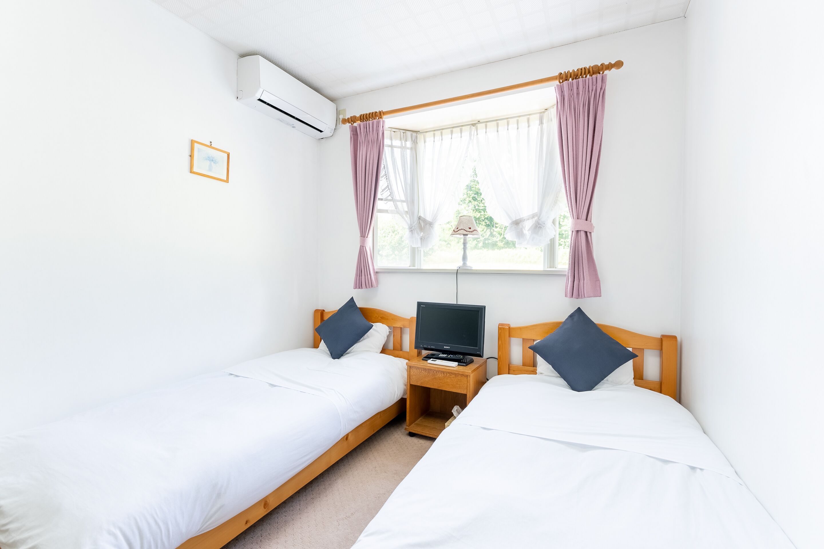 Ein Schlafzimmer mit zwei Einzelbetten, einem Nachttisch aus Holz mit Fernseher und einem Fenster mit Vorhängen.
