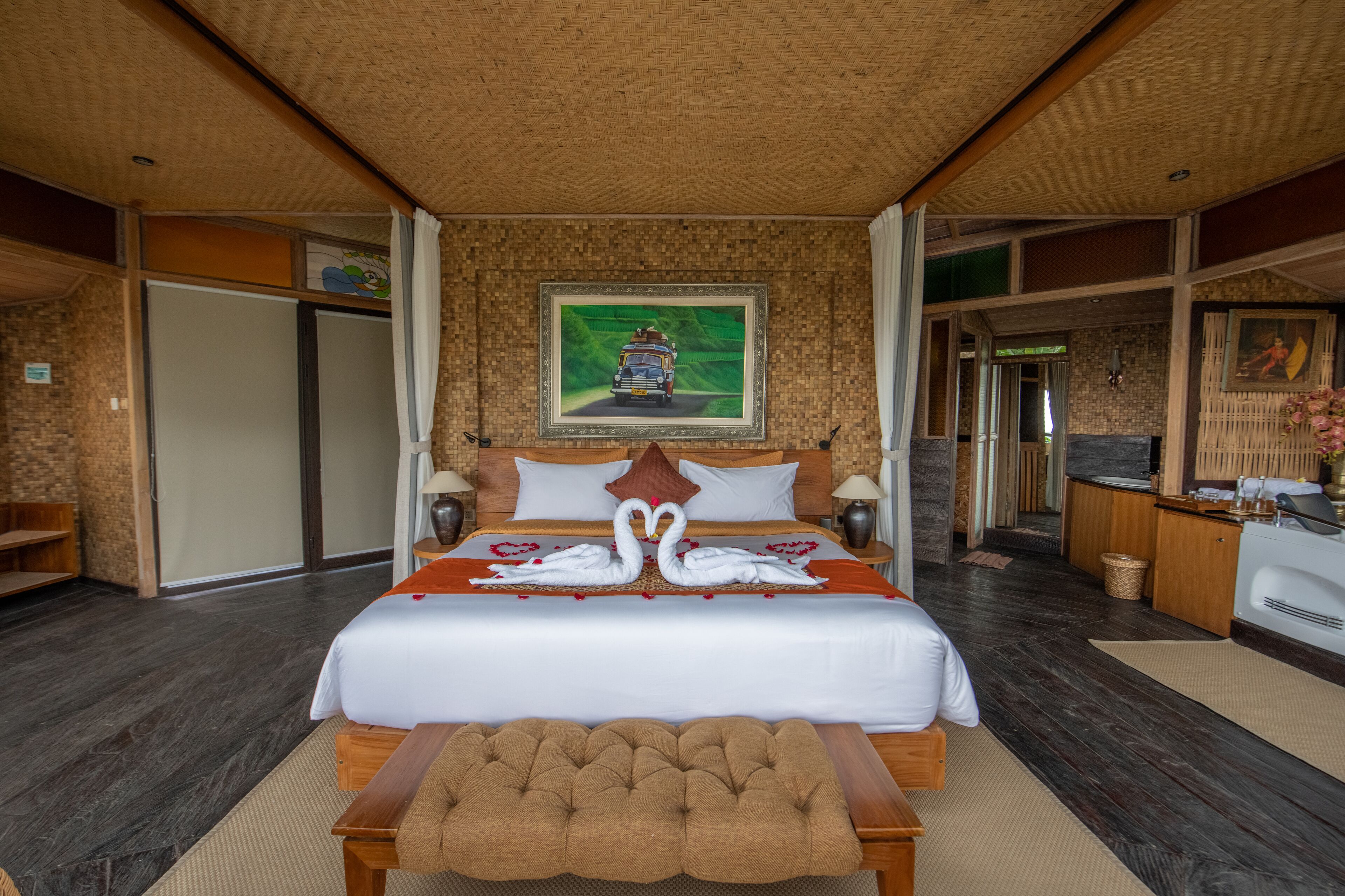 A hotel room with a large bed, a wooden bench, a painting on the wall, and a bathroom visible through an open door.