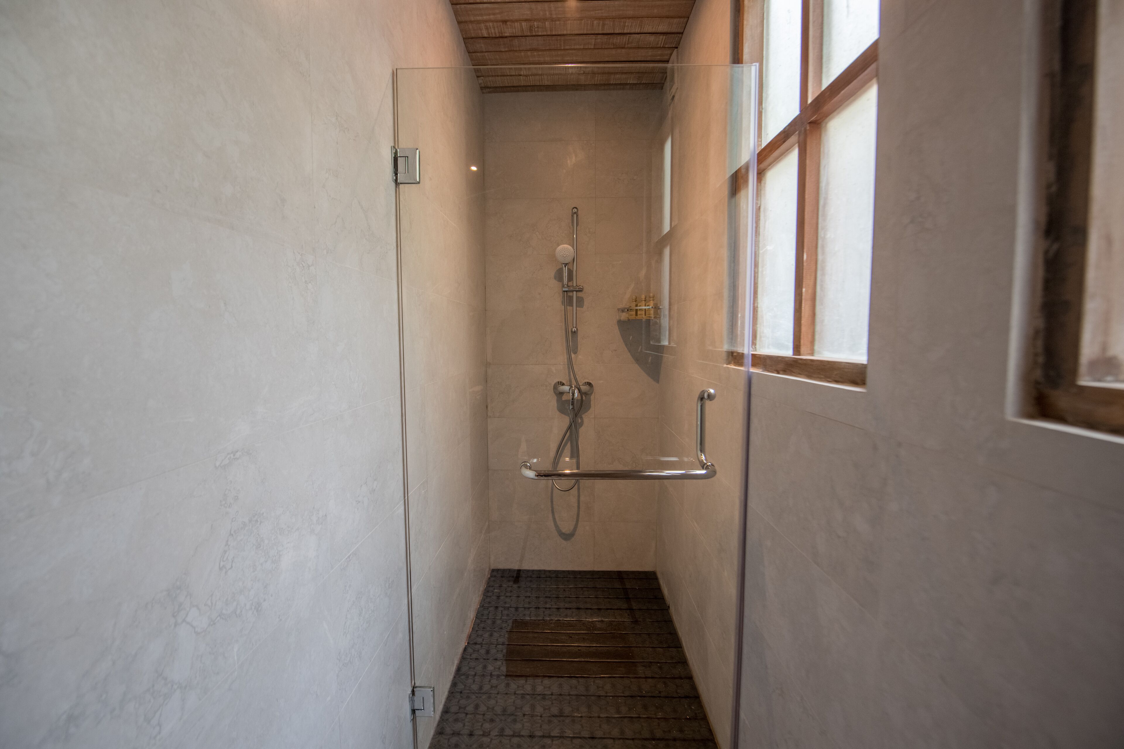 Un área de ducha estrecha con azulejos, una puerta de cristal, un cabezal de ducha y una barandilla.