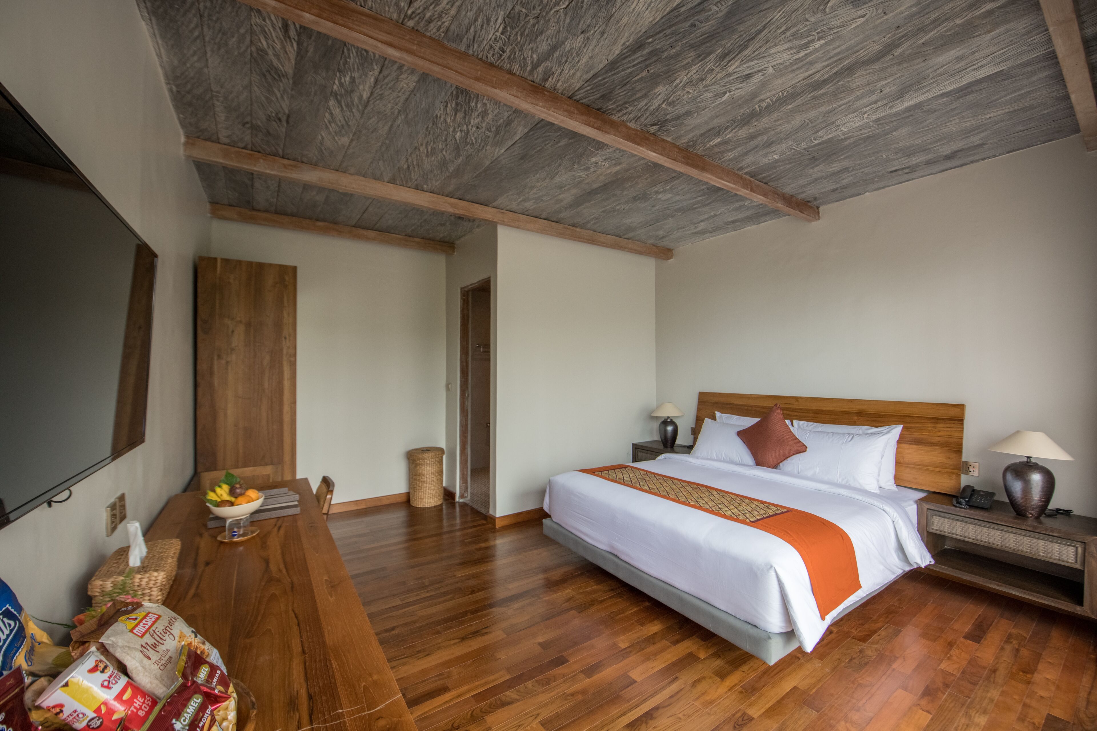 A hotel room with a wooden ceiling, a large bed with white and orange bedding, a wooden nightstand, a lamp, and a bowl of fruit on the table.