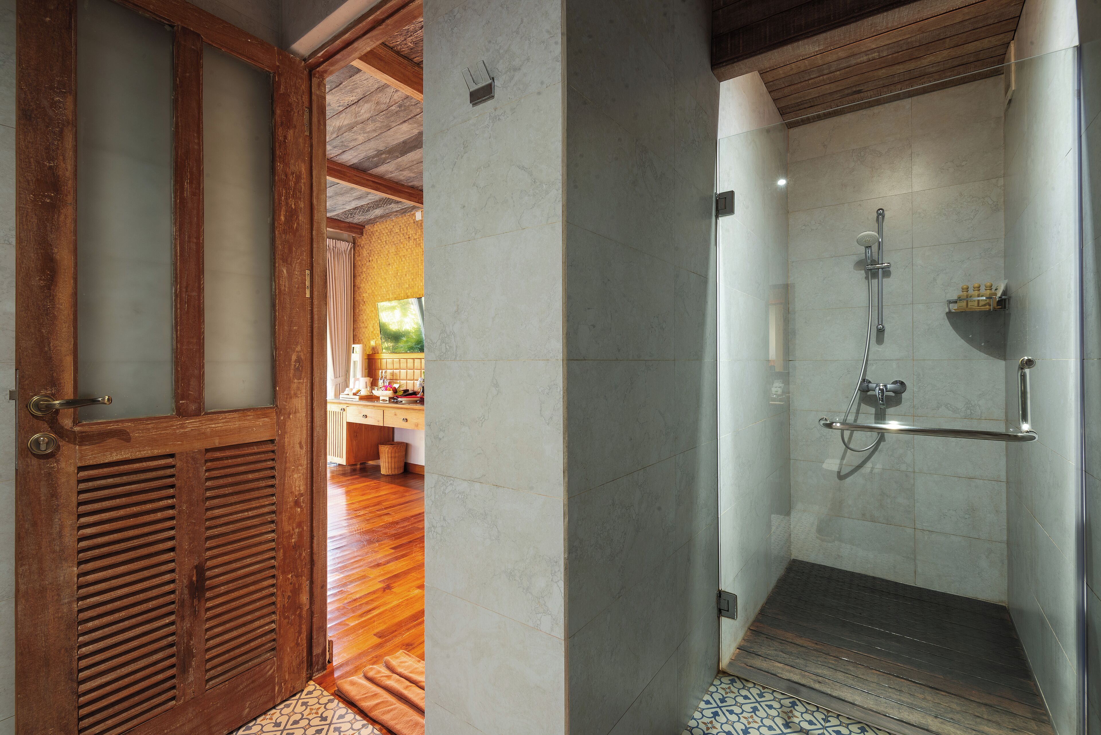 A bathroom with a shower, wooden door, and a view into a room with a wooden floor and a window.