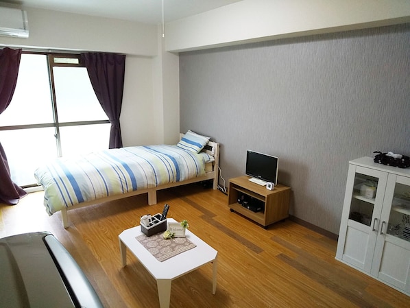 A small, neatly arranged room with a bed, a TV on a stand, a small table with a welcome sign, and a cabinet.
