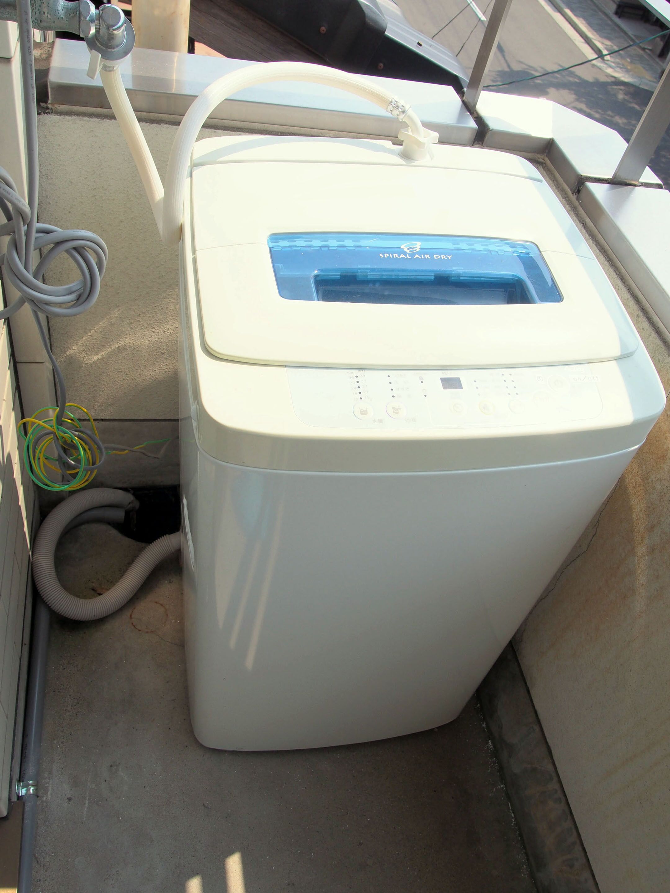 A compact, white, rectangular air conditioning unit with a blue control panel, connected to a flexible hose and a coiled cable.