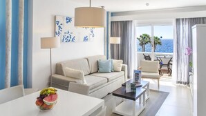 Ein modernes Hotelzimmer mit einer Couch, einem Esstisch und einer Obstschale. Ein Balkon bietet Blick auf das Meer und Palmen.