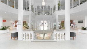 Ein geräumiges, mehrgeschossiges Atrium mit weißen Säulen, einer zentralen Treppe und einem Kronleuchter.