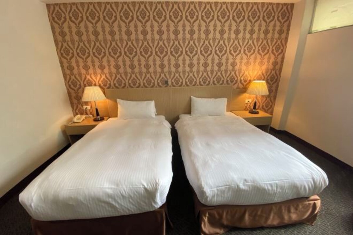 A hotel room with two single beds, a patterned headboard, bedside lamps, and a carpeted floor.