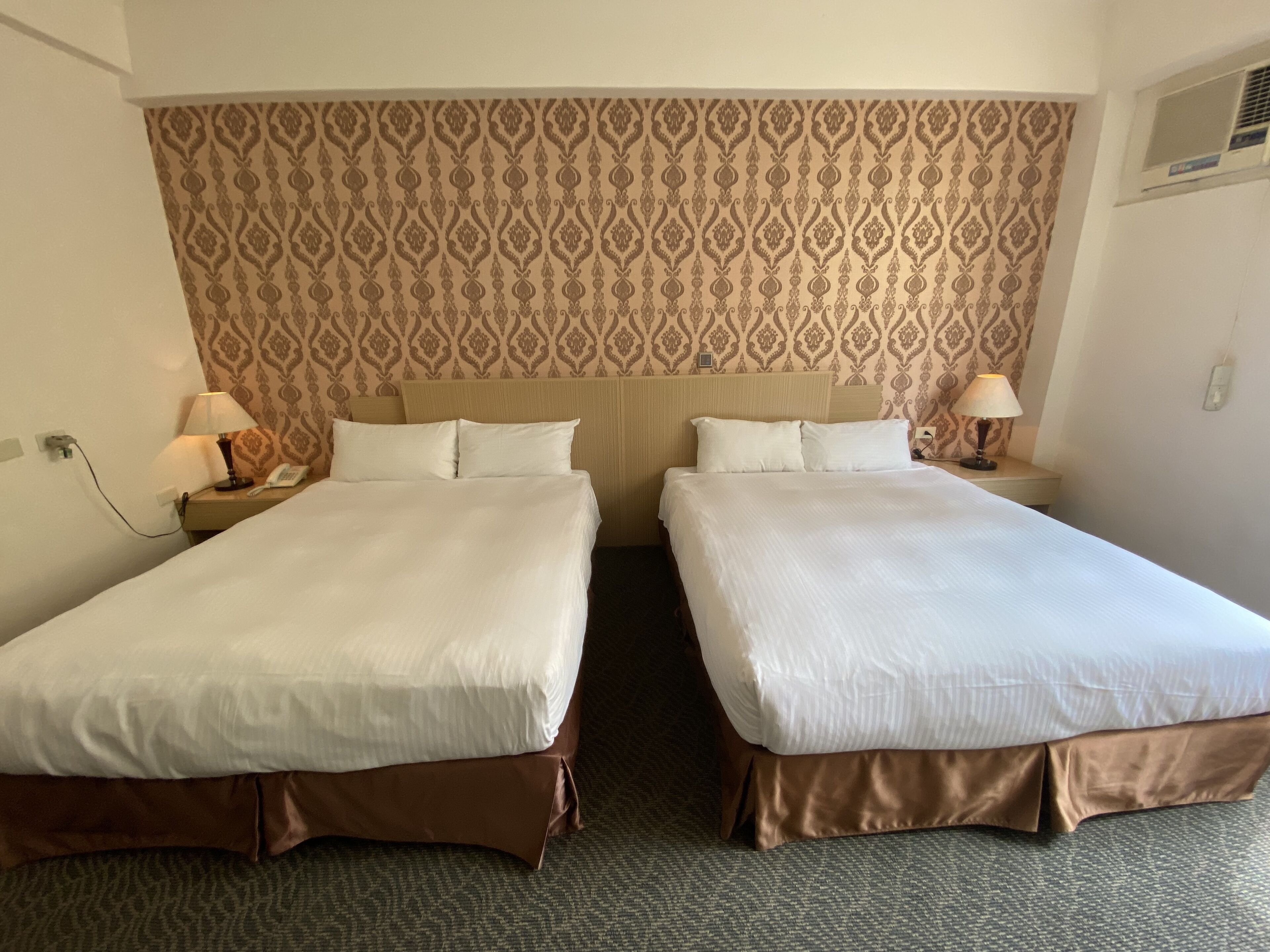 A hotel room with two beds, a patterned wallpaper, bedside lamps, and a carpeted floor.