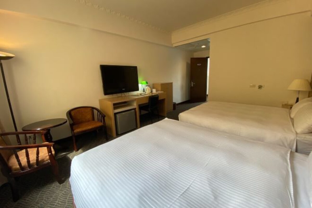 A hotel room with two beds, a desk with a television, a chair, and a lamp.