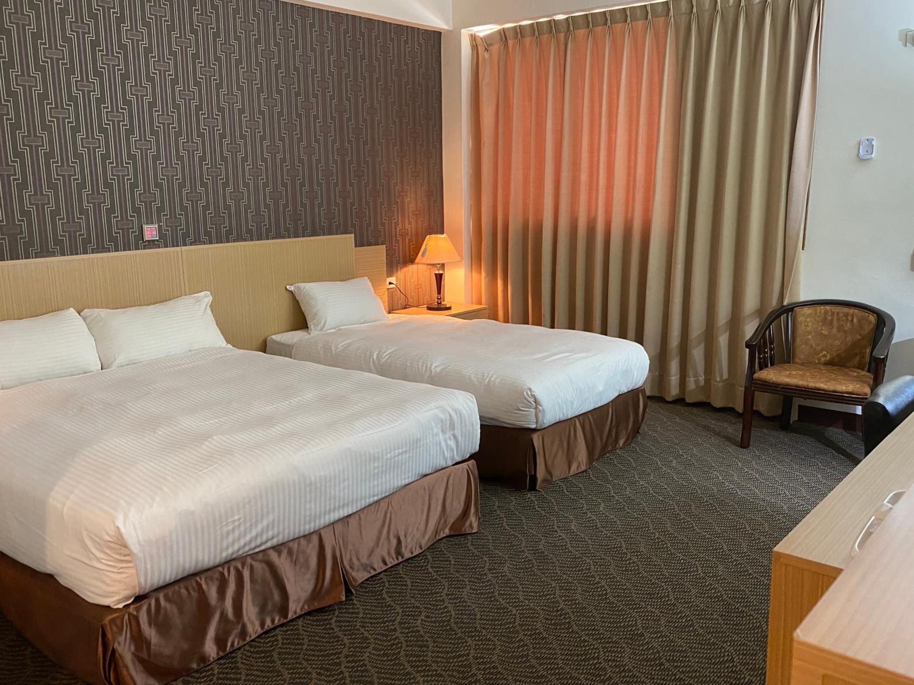 A hotel room with two beds, a chair, a lamp, and a patterned wall.