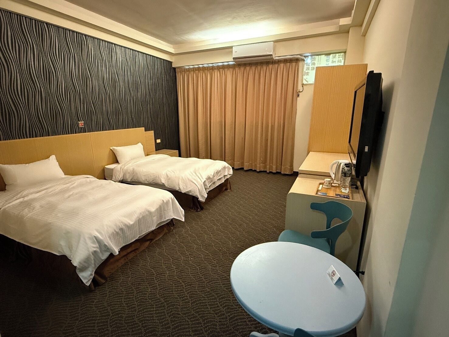 A hotel room with two beds, a desk with a chair, a small table, an air conditioning unit, and a television.