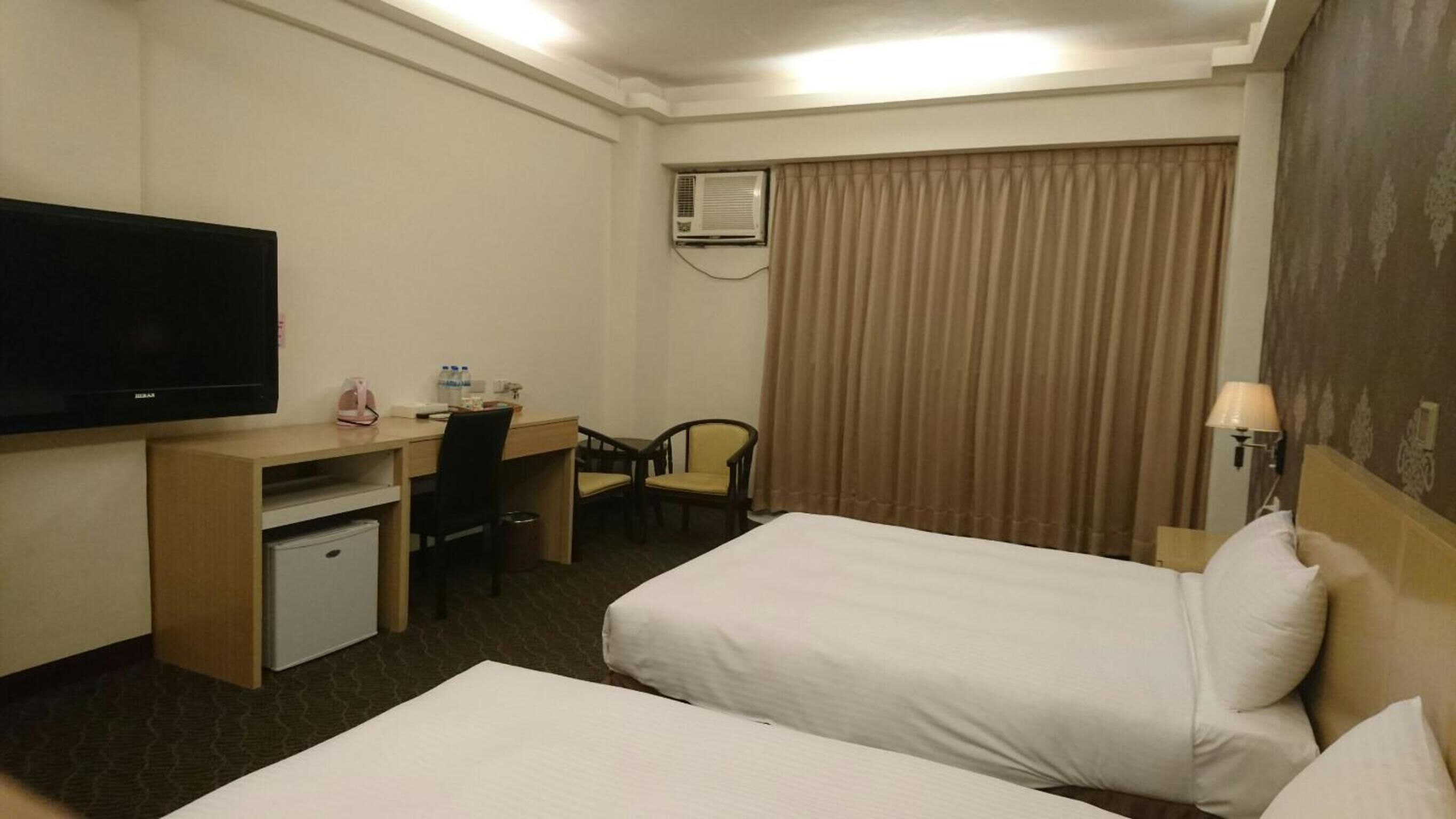 A hotel room with two beds, a desk with a chair, a television, an air conditioning unit, and a lamp.
