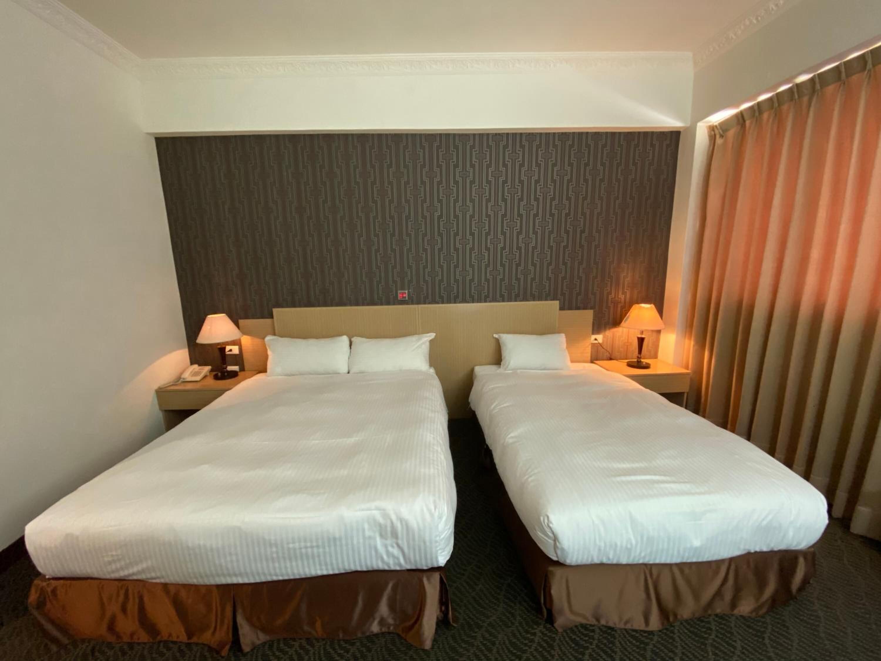 Habitación de hotel con dos camas individuales, cabecera con diseño estampado, lámparas de noche y una cortina a la derecha.