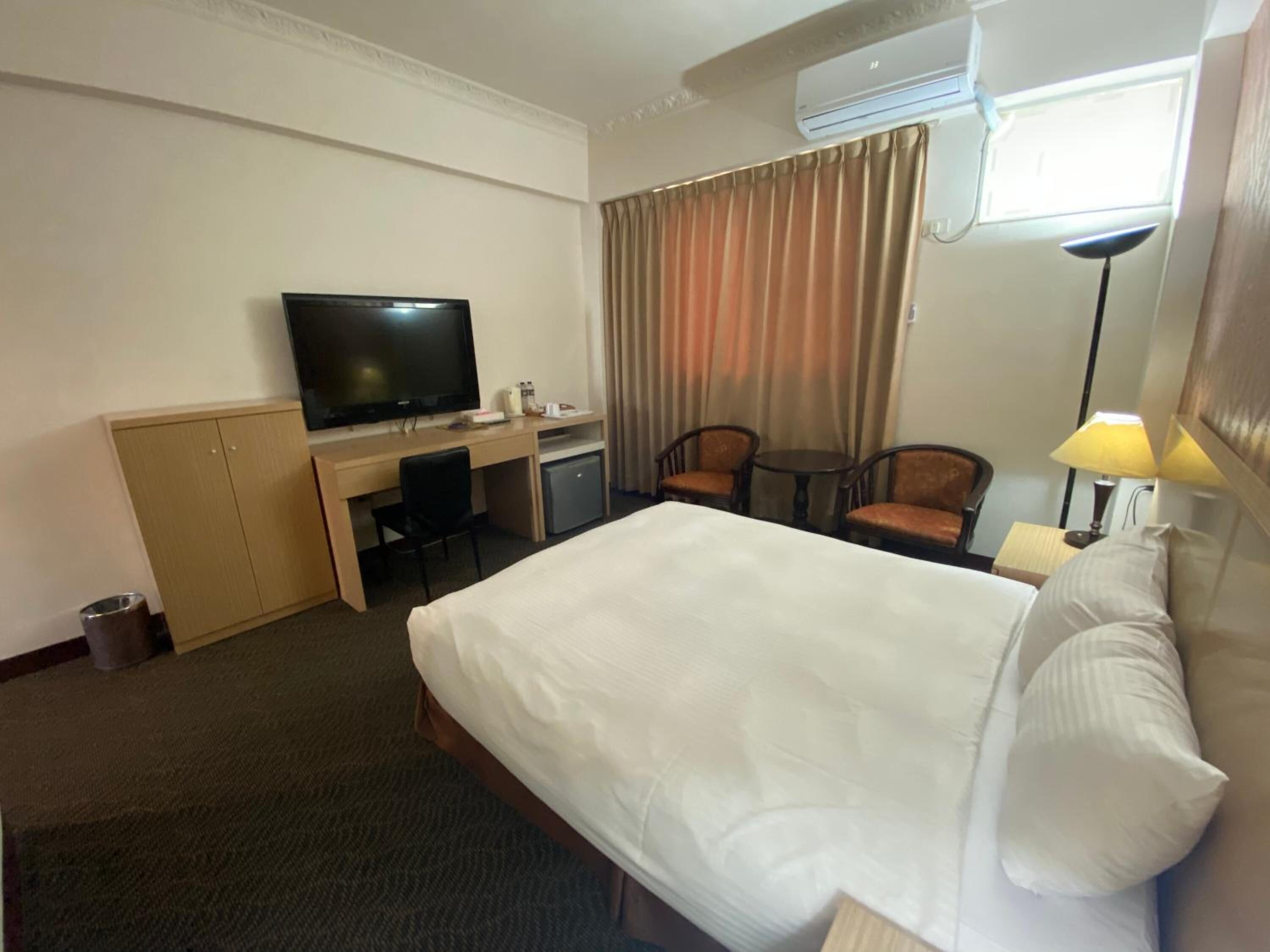 A hotel room with a bed, a desk with a television, a small refrigerator, two chairs, a lamp, and a window with curtains.