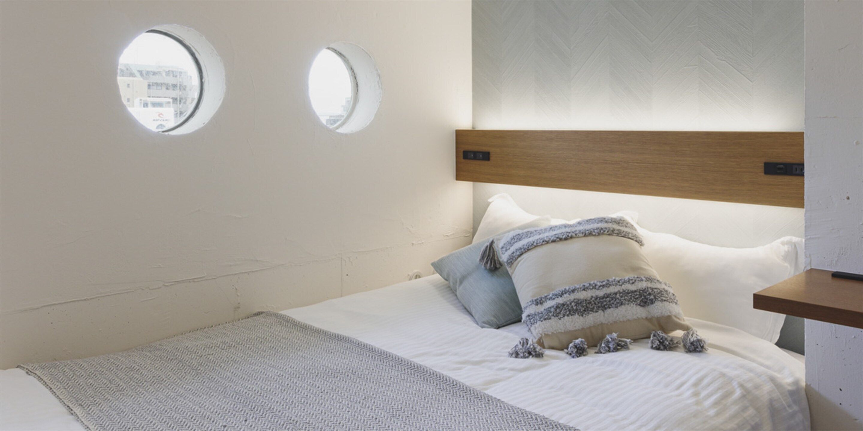 A bedroom with a bed, two round windows, a wooden headboard, and a small wooden shelf.