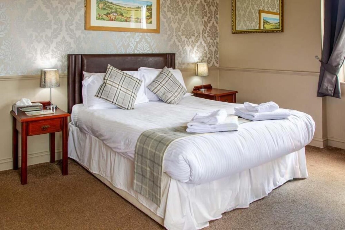 A neatly made bed with white linens and a plaid blanket, two bedside tables with lamps, and a framed landscape painting on the wall.