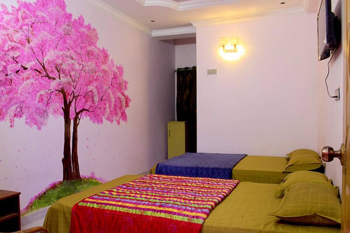 A bedroom with two beds, a wall mural of a pink tree, and a bedside lamp.