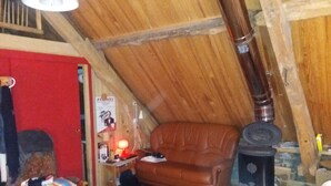 A cozy attic room with a wooden sloped ceiling, a brown leather sofa, a small wooden table with a lamp, and a vintage stove. - BRIGHT, SPACIOUS, COSY BARN AT THE FOOT OF THE CHEMIN DE SAINT-JACQUES (GAVARNIE GEDRE)