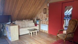 A cozy attic room with a wooden ceiling, a red door, a white radiator, a beige sofa, a glass coffee table, and a wooden chair. - BRIGHT, SPACIOUS, COSY BARN AT THE FOOT OF THE CHEMIN DE SAINT-JACQUES (GAVARNIE GEDRE)