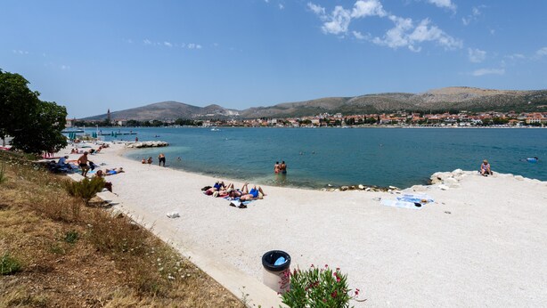 Una playa con aguas azules y transparentes, gente tomando el sol y un pueblo a lo lejos.