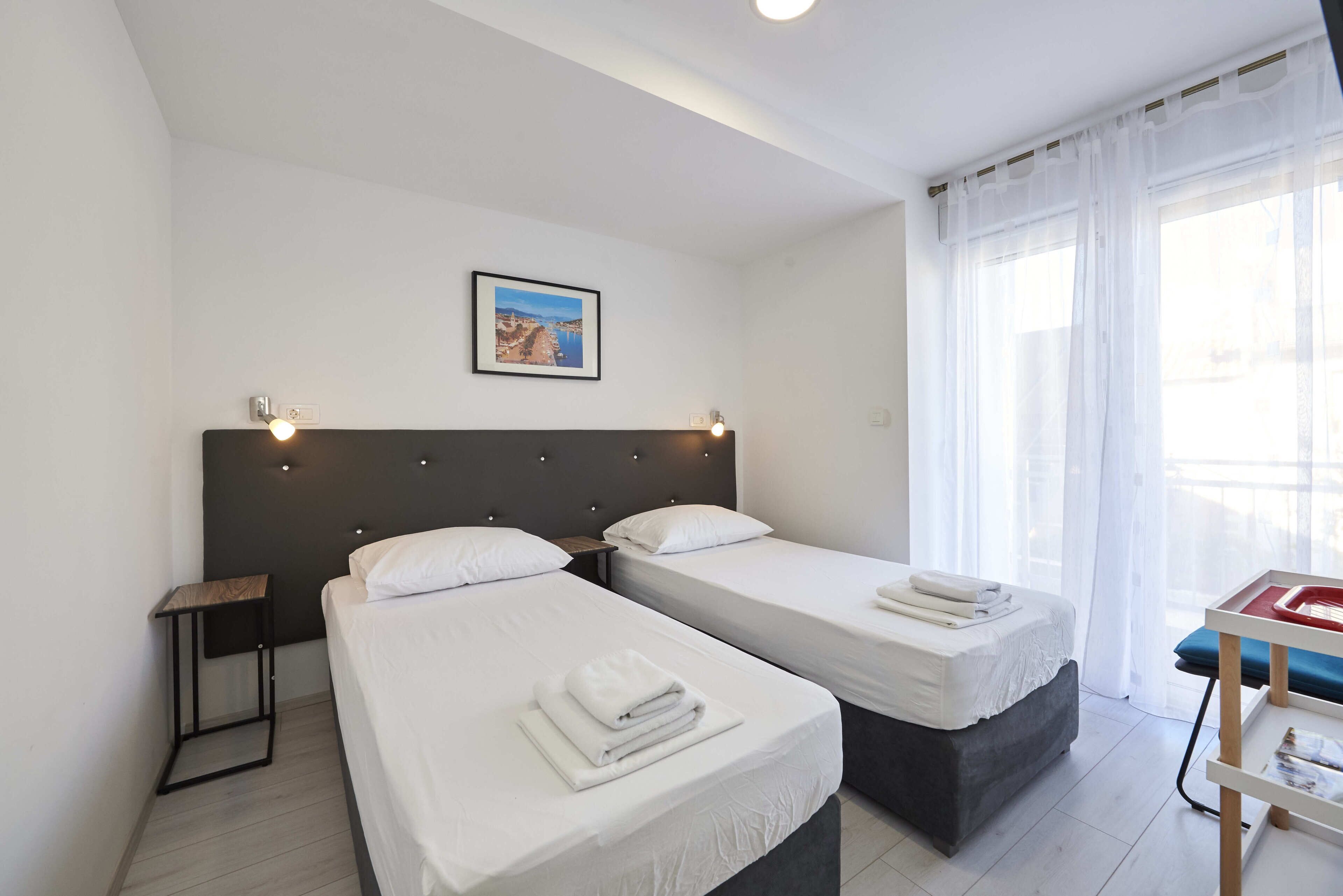 Habitación de hotel con dos camas, una mesita de noche, un cuadro enmarcado en la pared y una ventana con cortinas traslúcidas.