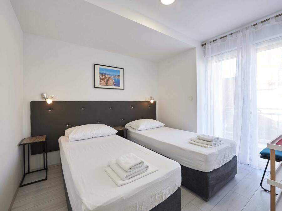 Habitación de hotel con dos camas, una mesita de noche, un cuadro enmarcado en la pared y una ventana con cortinas traslúcidas.