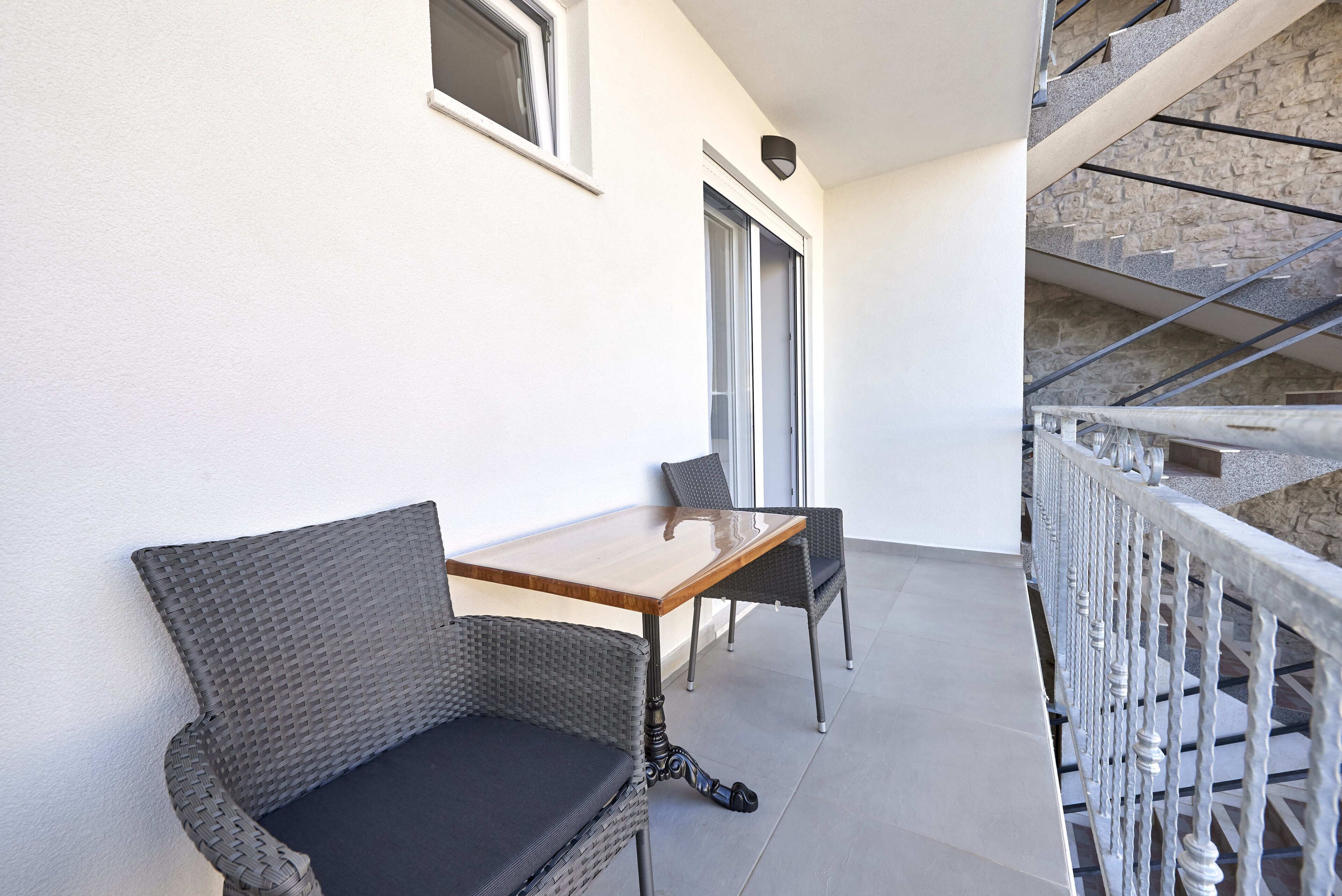 A balcony with a wooden table and two wicker chairs, a stone wall, and a metal railing.