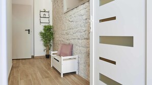 Un pasillo con pared de piedra, una puerta blanca con espejo, un estante pequeño con una planta y un banco blanco con cojín.