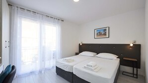 Una cama doble con sábanas blancas, una mesita de noche con lámpara y una ventana con cortinas traslúcidas.
