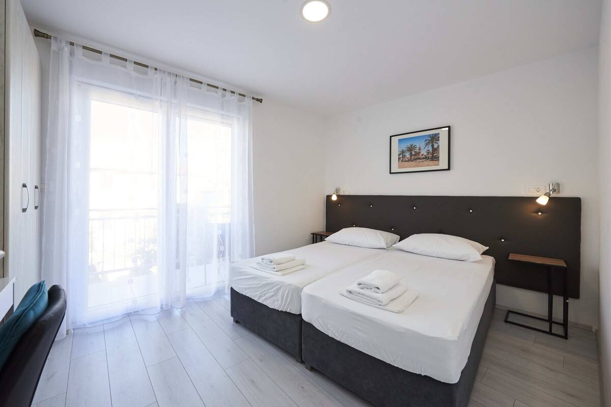 Ein Doppelbett mit weißen Laken, ein Nachttisch mit Lampe und ein Fenster mit transparenten Vorhängen.
