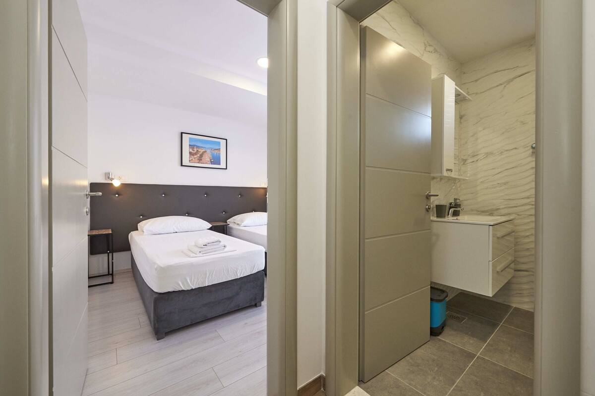 Ein modernes Hotelzimmer mit zwei Betten, einem Badezimmer mit Marmorwand und einem kleinen Nachttisch mit Lampe.