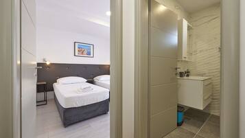 A modern hotel room with two beds, a bathroom with a marble wall, and a small bedside table with a lamp.