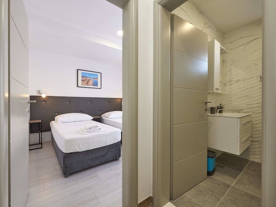 A modern hotel room with two beds, a bathroom with a marble wall, and a small bedside table with a lamp.
