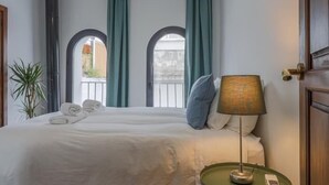 Hotellihuone, jossa on sänky, yöpöytä lampun kanssa ja kaksi ikkunaa, joissa on verhot.