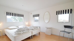 Ein schlicht eingerichtetes Schlafzimmer mit einem Bett, einem Sessel, einem runden Spiegel und zwei Fenstern mit schwarz-weiß gestreiften Vorhängen.