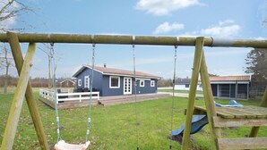 Ein hölzernes Schaukelgestell, eine Autoreifen-Schaukel und ein blaues Haus mit einer Terrasse in einem grünen Garten.