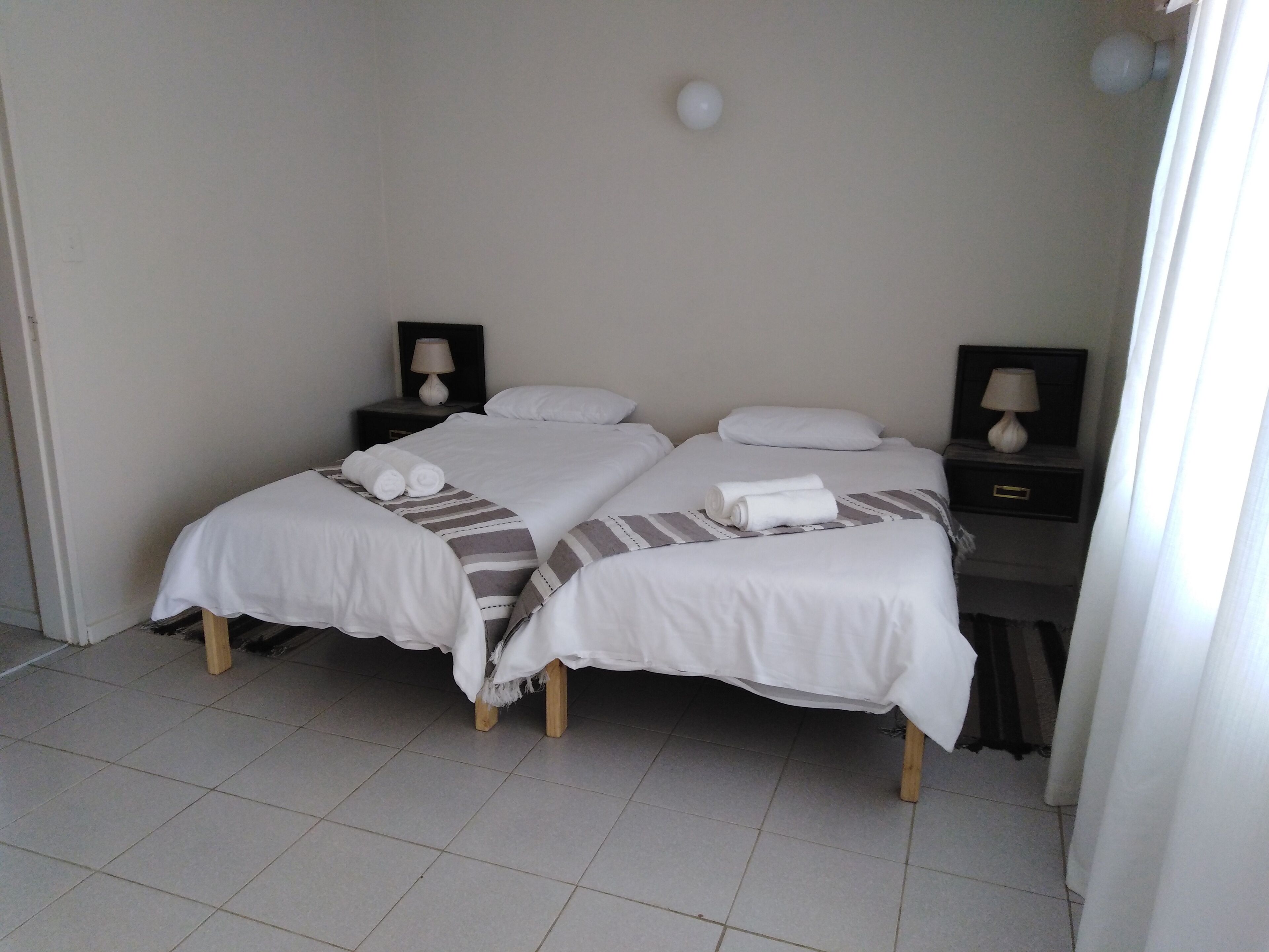 Ein Hotelzimmer mit zwei Betten, jeweils mit einem Kopfkissen und einem aufgerollten Handtuch. An den Nachttischen stehen Lampen und eine kleine Schublade.
