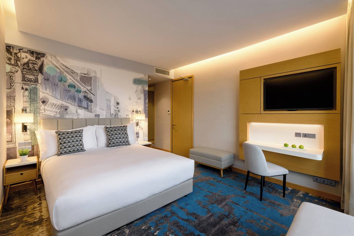 A modern hotel room with a large bed, a desk with a flat-screen TV, a small seating area, and a cityscape mural on the wall.