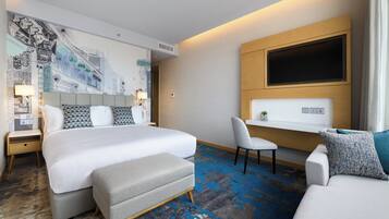 A modern hotel room with a large bed, a desk, a sofa, and a mural on the wall.