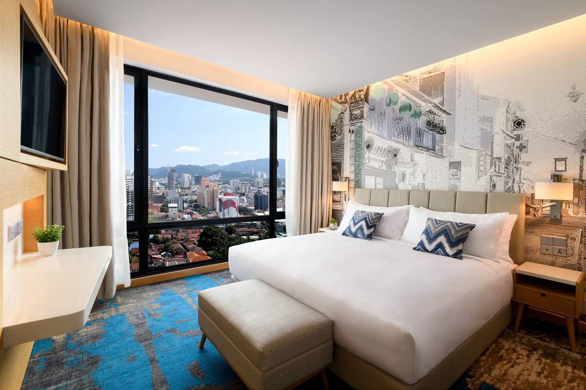 A hotel room with a large bed, a desk, and a view of a cityscape through a large window.
