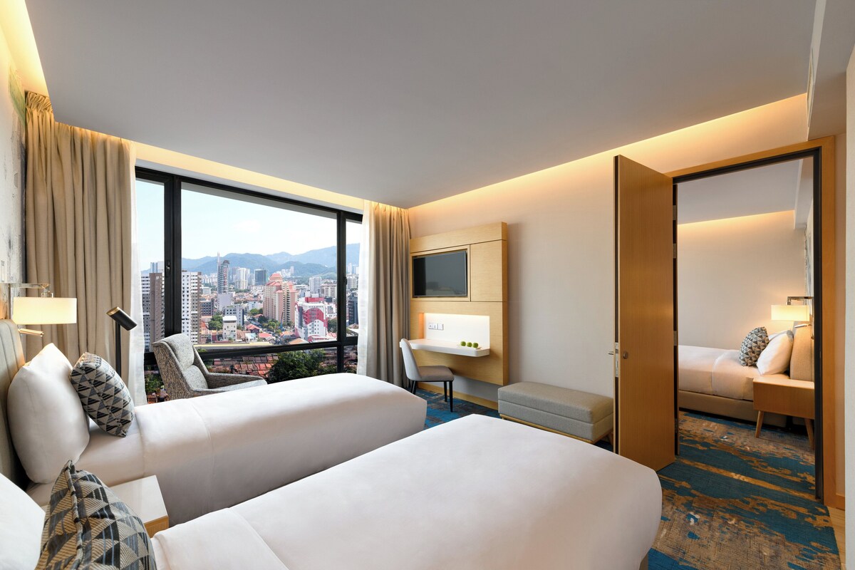 A hotel room with two beds, a desk, a chair, a TV, and a view of the city.