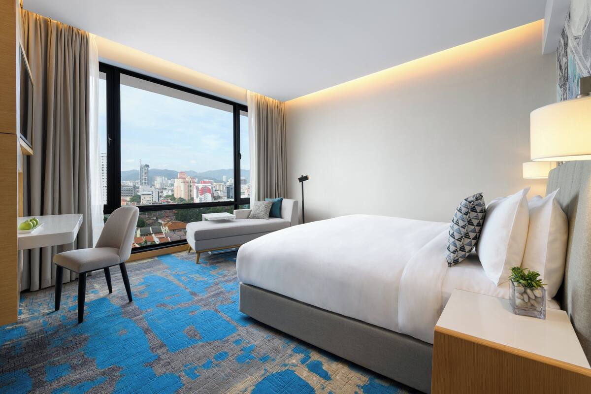 A modern hotel room with a large bed, a desk, a chair, and a view of the cityscape through large windows.