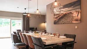 Een eetruimte met een grote tafel voor zes personen, omringd door lederen stoelen. Boven de tafel hangen hanglampen en aan de muur hangt een schilderij met een strandthema.