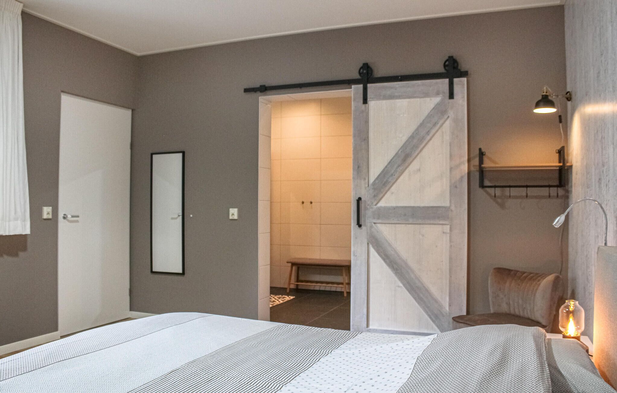 Een slaapkamer met een bed, een schuifdeur naar een badkamer, een nachtkastje met een lamp en een wandplank.