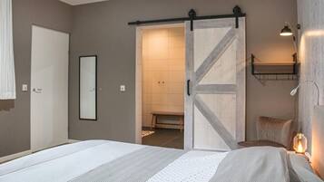 Een slaapkamer met een bed, een schuifdeur naar een badkamer, een nachtkastje met een lamp en een wandplank.