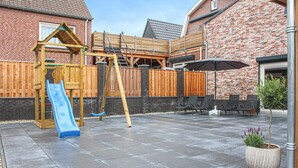 Een achtertuin met een houten speeltoestel, een glijbaan en een zithoek met stoelen en een parasol.