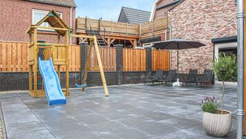 Een achtertuin met een houten speeltoestel, een glijbaan en een zithoek met stoelen en een parasol.