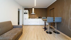 Una cocina moderna con armarios blancos, horno empotrado y campana extractora de acero inoxidable. Hay una zona de comedor con una mesa de madera y dos taburetes de bar azules.